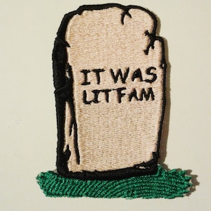 Puede incluir: Parche bordado con forma de lápida beige con el texto negro "IT WAS LIT FAM". La lápida tiene un borde negro y está sobre un parche de hierba verde.