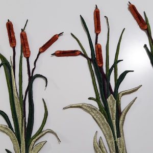 May include: A set of three embroidered iron-on patches featuring a cluster of brown cattail reeds with green leaves.