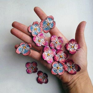 Op de afbeelding: Een verzameling geborduurde bloemenpatches in tinten roze, blauw en wit. De patches zijn op een hand gerangschikt, waardoor hun delicate details worden getoond.