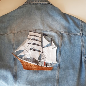 May include: A blue denim jacket with a white embroidered sailboat on the back. The sailboat has a brown hull and white sails.