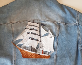 May include: A blue denim jacket with a white embroidered sailboat on the back. The sailboat has a brown hull and white sails.