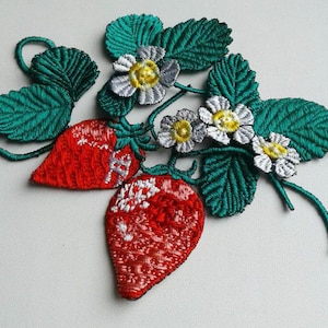 Puede incluir: Parche de fresas bordadas con dos fresas rojas, detalles blancos, hojas verdes y pequeñas flores blancas y amarillas. El parche está hecho con costuras detalladas, mostrando un diseño vibrante y texturizado.