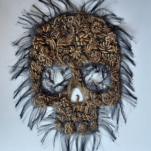 May include: A black and gold skull embroidered with floral patterns. The skull is surrounded by black feathers.