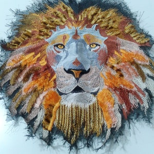 May include: A close-up of an embroidered lion's face with a black, orange, and gold mane. The lion's eyes are a light blue colour and the background is a white surface.