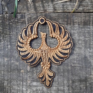 May include: A gold embroidered patch depicting a stylised bird with outstretched wings. The bird is outlined in black and features a detailed design.