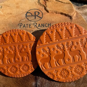 K&ouml;nnte beinhalten: Zwei runde, braune Lederuntersetzer mit festlichem Design. Jeder Untersetzer zeigt gepr&auml;gte Rentiere, Weihnachtsb&auml;ume und Schneeflockenmuster. Die Untersetzer sind von Pate Ranch, Est. 2015.