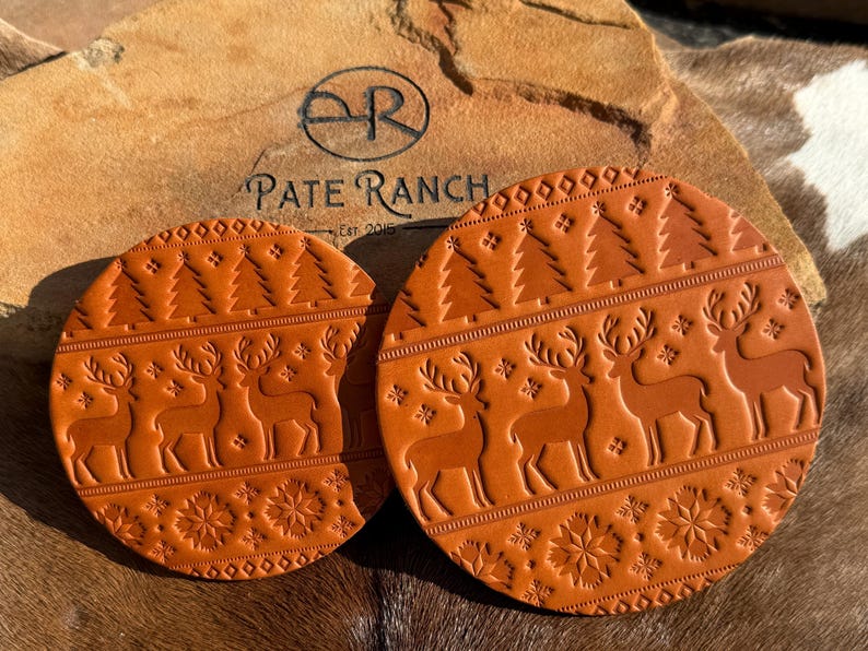 K&ouml;nnte beinhalten: Zwei runde, braune Lederuntersetzer mit festlichem Design. Jeder Untersetzer zeigt gepr&auml;gte Rentiere, Weihnachtsb&auml;ume und Schneeflockenmuster. Die Untersetzer sind von Pate Ranch, Est. 2015.