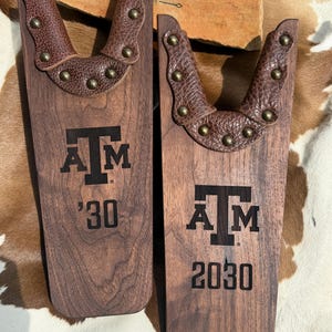 May include: Two wooden boot jacks with brown leather accents and brass studs. Each jack features the letters "A M" and a year, one with "'30" and the other with "2030". The background includes a cowhide pattern.