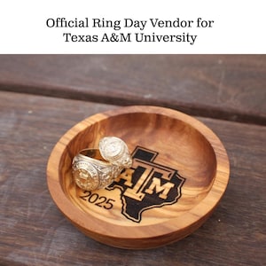 Puede incluir: Un cuenco de madera con dos anillos dorados en su interior. El cuenco tiene el logotipo de la Universidad de Texas A&M y el texto "2025" grabado en la madera. El texto "Official Ring Day Vendor for Texas A&M University" está en la parte superior.