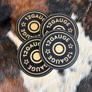 May include: Four black leather coasters with gold stitching and the text "12 GAUGE" in gold lettering. The coasters are stacked on top of each other.