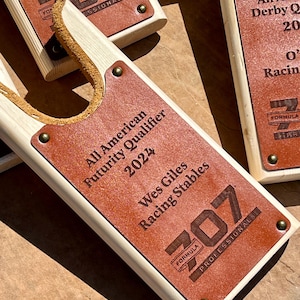 May include: A wooden horse stall plaque with a brown leather patch. The leather patch has the text "All American Futurity Qualifier 2024 Wes Giles Racing Stables" and the number "707" with the text "FORMULA PROFESSIONAL".