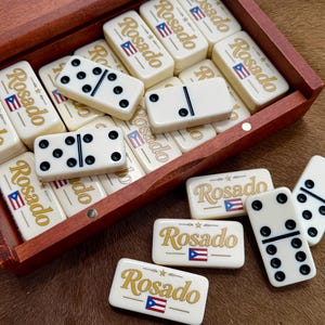 May include: A set of off-white dominoes with black pips, gold text reading Rosado, and a small Puerto Rican flag graphic. The dominoes are stored in a rectangular wooden box with a hinged lid, with several pieces resting outside the container.