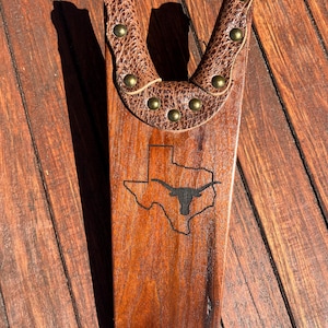 May include: A wooden boot jack with a brown leather yoke secured with brass rivets. The jack features a carved outline of the state of Texas with a longhorn silhouette.