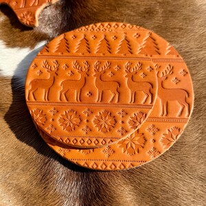 K&ouml;nnte beinhalten: Zwei runde, orangebraune Lederuntersetzer mit gepr&auml;gten Feiertagsmotiven. Ein Untersetzer zeigt eine Reihe von Rentieren und Weihnachtsb&auml;umen, w&auml;hrend der andere Schneeflockenmuster hat. Zus&auml;tzliche Lederausschnitte mit &auml;hnlichen Designs sind ebenfalls sichtbar.