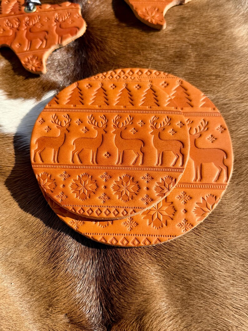 K&ouml;nnte beinhalten: Zwei runde, orangebraune Lederuntersetzer mit gepr&auml;gten Feiertagsmotiven. Ein Untersetzer zeigt eine Reihe von Rentieren und Weihnachtsb&auml;umen, w&auml;hrend der andere Schneeflockenmuster hat. Zus&auml;tzliche Lederausschnitte mit &auml;hnlichen Designs sind ebenfalls sichtbar.