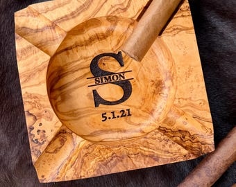 May include: A square wooden ashtray with a natural grain pattern, featuring a central engraving of the letter S, the name SIMON, and the date 5.1.21. A brown cigar with a label reading MEYER rests across one of the four corner notches.
