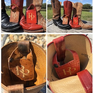 May include: A collection of cowboy boots in various colors, including black, red, and brown. Each boot has a leather tag with the initials "RP" in gold lettering. The boots are displayed outdoors, suggesting a Western or country theme.