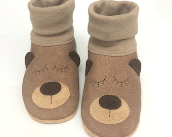 May include: A pair of brown leather baby booties with a bear face design. The booties have a beige cuff and are made of soft, comfortable material.