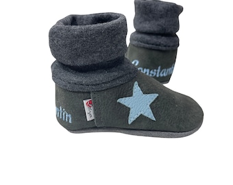 Puede incluir: Un par de botines de bebé gris oscuro con una estrella azul claro en la parte delantera. Los botines están hechos de cuero suave y tienen un puño de punto gris. Los botines están personalizados con los nombres "Martin" y "Constantin".