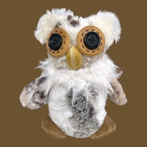 May include: A plush owl toy featuring long, shaggy white and gray fur. The eyes are constructed from large, tan circular buttons with black centers. A small, vertically stitched golden-yellow thread beak sits between the eyes. Two flat, tan fabric feet are visible at the base.