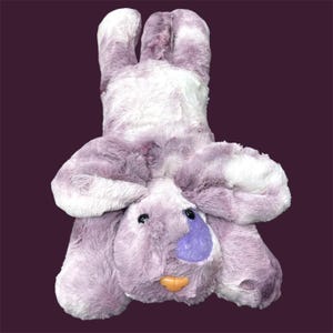 May include: A plush bunny toy in shades of purple and white. The bunny has long ears, a purple nose, and an orange mouth. The toy is made of soft, fluffy material and is designed to be cuddled.