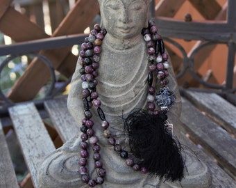 May include: A gray stone Buddha statue wearing a long beaded necklace with black and pink beads and a black tassel. The necklace has a silver charm with a geometric design.