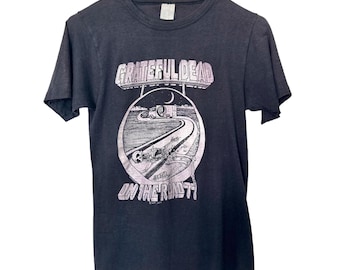 May include: Dark gray t-shirt featuring the words "Grateful Dead" above a circular graphic with a road, skeleton, and the text "On the Road". The shirt is hanging on a white hanger.