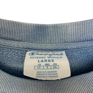 Pode incluir: Close-up de um moletom Champion Reverse Weave azul claro. A etiqueta diz "LARGE" e inclui informa&ccedil;&otilde;es de tamanho para CA, MEX e JPN. O moletom &eacute; feito em El Salvador.