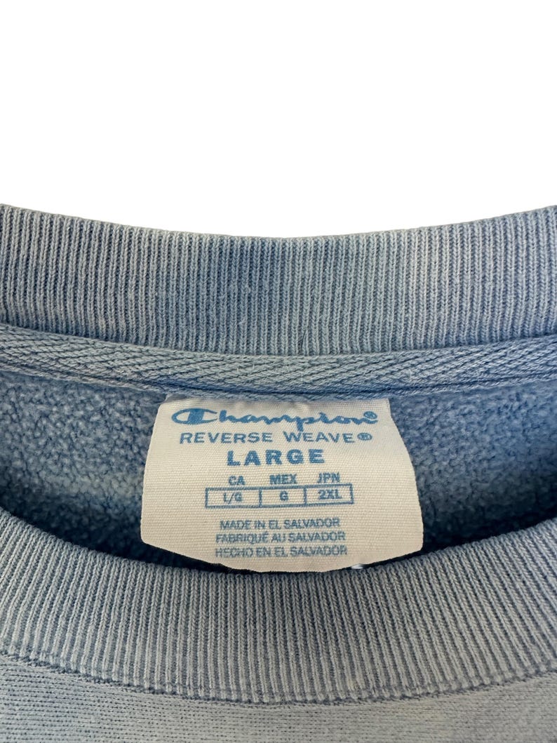 Pode incluir: Close-up de um moletom Champion Reverse Weave azul claro. A etiqueta diz "LARGE" e inclui informa&ccedil;&otilde;es de tamanho para CA, MEX e JPN. O moletom &eacute; feito em El Salvador.