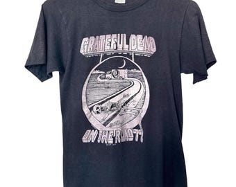 May include: Dark grey t-shirt with a graphic featuring the words "Grateful Dead" above a circular image of a road, a truck, and the words "On The Road '77". The shirt is hanging on a white hanger.