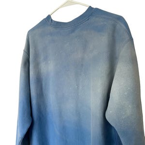 Pode incluir: Um moletom Champion azul claro com efeito degrad&ecirc;. O moletom de gola redonda est&aacute; pendurado em um cabide branco. As mangas s&atilde;o compridas e o tecido parece ser uma mistura macia de algod&atilde;o.