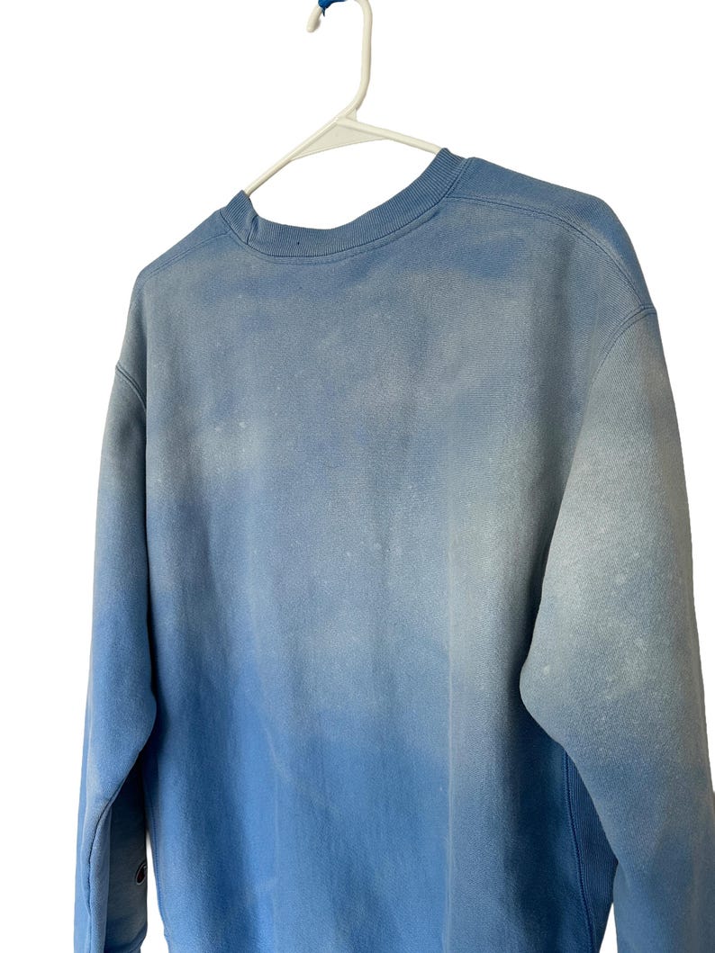 Pode incluir: Um moletom Champion azul claro com efeito degrad&ecirc;. O moletom de gola redonda est&aacute; pendurado em um cabide branco. As mangas s&atilde;o compridas e o tecido parece ser uma mistura macia de algod&atilde;o.