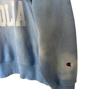 Pode incluir: Camisola Champion azul claro com letras brancas. A manga apresenta o emblema do log&oacute;tipo Champion. O tecido tem um aspeto desbotado e vintage. Uma pe&ccedil;a cl&aacute;ssica de roupa casual.