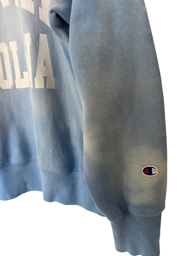 Pode incluir: Camisola Champion azul claro com letras brancas. A manga apresenta o emblema do log&oacute;tipo Champion. O tecido tem um aspeto desbotado e vintage. Uma pe&ccedil;a cl&aacute;ssica de roupa casual.