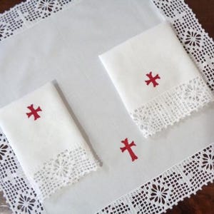 May include: White linen set with red cross embroidery. The set includes a large square cloth with a decorative crocheted lace border, and two folded hand towels with a red cross and lace trim.