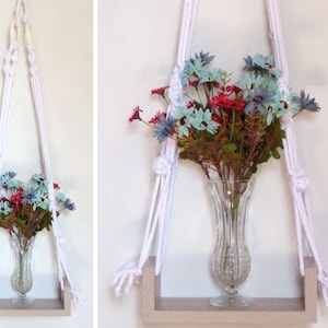 Könnte beinhalten: Ein weißer Makramee-Pflanzenhänger mit einem Holzregal, in dem eine klare Glasvase mit blauen und roten Blumen steht.