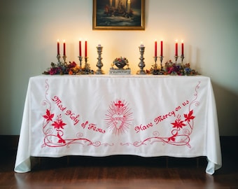 Può includere: Una tovaglia bianca con ricami rossi che raffigurano un cuore con una croce e le parole "Most Holy of Jesus" e "Have Mercy on us". La tovaglia è posta su un tavolo con candele accese e un dipinto incorniciato.