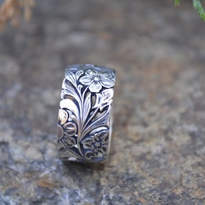 Könnte beinhalten: Ein silberner Ring mit einem floralen Design. Der Ring ist breit und hat ein detailliertes Design aus Blumen und Blättern.