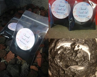 Op de afbeelding: Drie kleine, doorzichtige plastic zakjes gevuld met zwart houtskoolpoeder. Elk zakje heeft een wit rond etiket met de tekst "Charcoal Facial Peel beauty organics".
