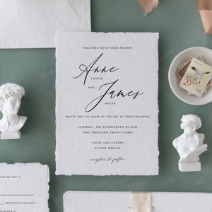 May include: Wedding invitation suite with white paper, a white envelope, and a white card with the text "Anne Cooper and James Miller" in black calligraphy. The invitation is decorated with white flowers, a white ribbon, and two white plaster busts.