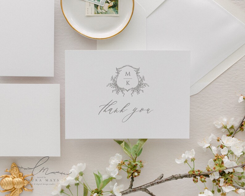 May include: A white thank you card with a grey monogram of the letters M and K in a shield with floral accents. The text "thank you" is written in cursive below the monogram.