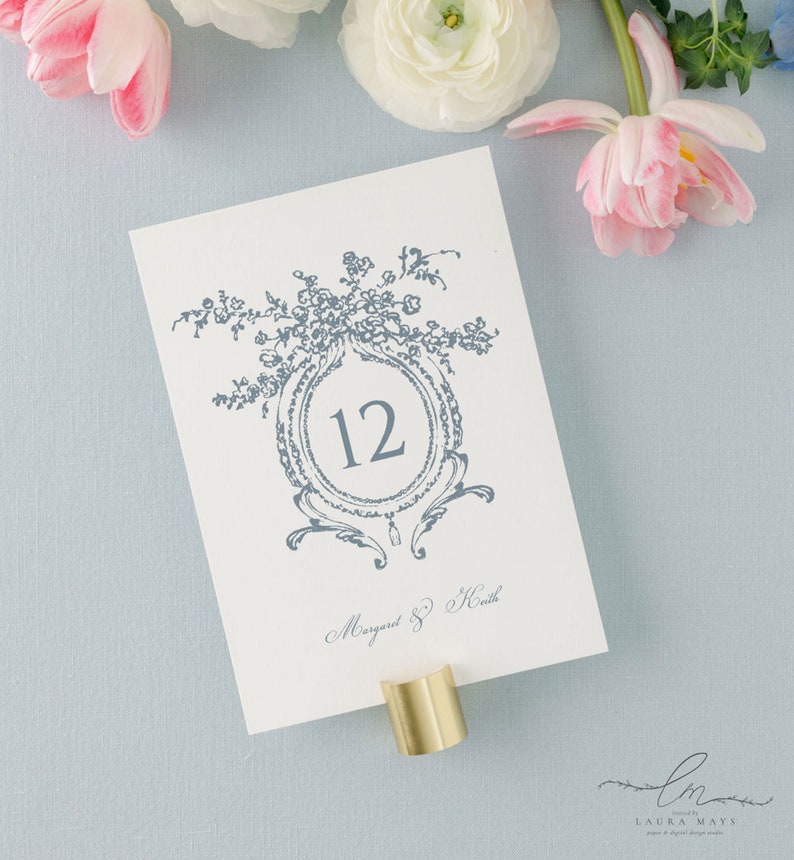 May include: A white card with a blue floral design and the number 12 in a circle. The text "Margaret & Heath" is written in cursive at the bottom of the card.
