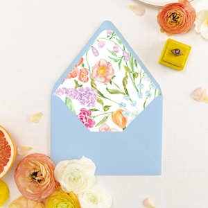 May include: A light blue envelope with a floral patterned interior. The floral design features pink, purple, and orange flowers with green leaves on a white background. The envelope is surrounded by flowers and a yellow ring box.