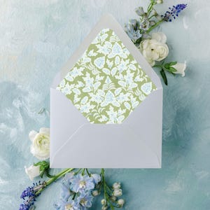 May include: A white envelope with a floral patterned interior, featuring a design of white and blue leaves on a green background. The envelope is surrounded by white and blue flowers, set against a light blue painted backdrop.