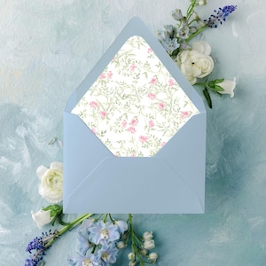 May include: A light blue envelope with a floral patterned interior, featuring pink roses and green leaves on a white background. The envelope is surrounded by white and blue flowers against a light blue painted background.