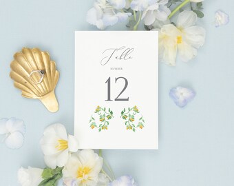 Puede incluir: Una tarjeta blanca con el texto "Table Number 12" en fuente gris. La tarjeta está decorada con dos pequeñas ilustraciones florales en verde y amarillo. La tarjeta está rodeada de flores blancas y azules sobre un fondo azul claro.