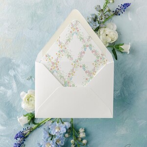 May include: A white envelope with a floral patterned liner, open to reveal the design. The liner features a delicate pattern of pink, yellow, and blue flowers. The envelope is surrounded by white and blue flowers on a light blue painted background.