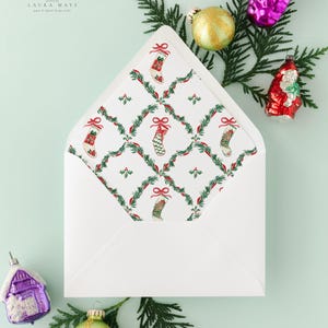 May include: A white envelope with a Christmas stocking pattern on the inside, surrounded by festive ornaments and evergreen branches. The envelope's interior features a repeating design of stockings, bows, and holly. The image includes the text "LAURA MAYS paper & digital design studio."