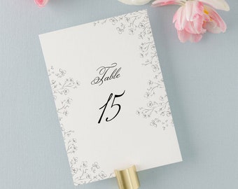 May include: A white table number card with the word "Table" and the number "15" in elegant black script. Delicate floral illustrations adorn the corners. The card is held by a gold-colored metal holder. Soft pink and white flowers are in the background.