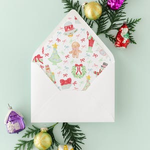 May include: A white envelope with a Christmas-themed patterned interior, surrounded by festive ornaments and evergreen sprigs. The interior design features illustrations of gingerbread men, Christmas trees, and bows. Ornaments include a purple house, a gold star, and a red Santa.